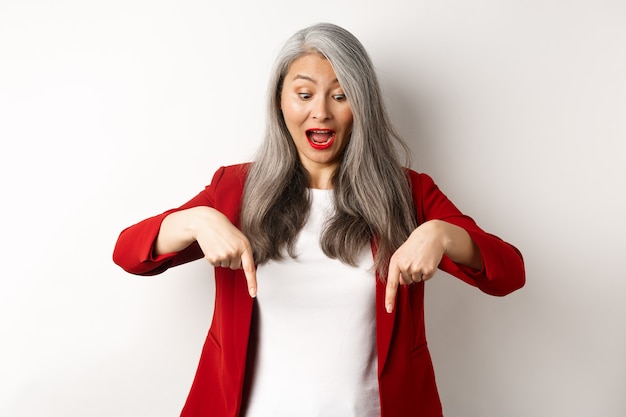 Stylish asian elderly woman in red blazer checking out special promotion, pointing fingers down and looking excited, scream of joy, white wall.