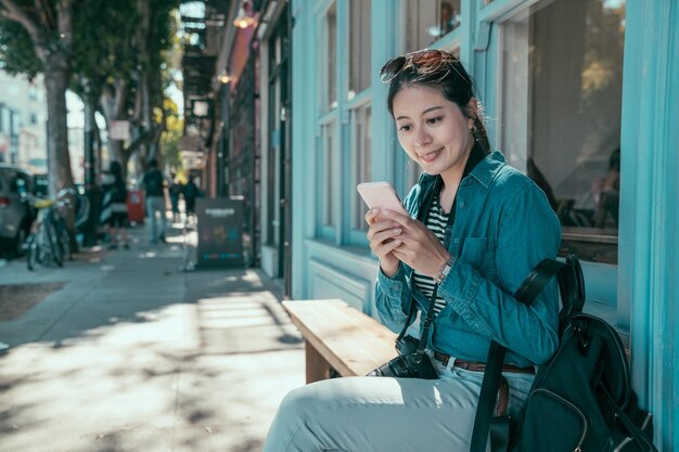 Stylish asian girl backpacker using apps on cellphone while\
sitting on bench on citywalk outside store during summer vacation.\
female blogger reading article on mobile phone relax on\
street.