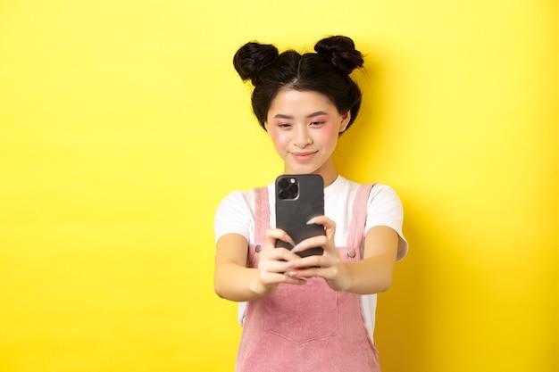 スタイリッシュなアジアの女の子がスマートフォンで写真を撮り、携帯電話でビデオを作り、笑顔で、黄色に立っています。