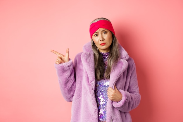 Стильная азиатская дама с седыми волосами, в модном пальто из искусственного меха и повязкой на голову, жалуется на что-то плохое, хмурится и указывает влево, стоит на розовом фоне