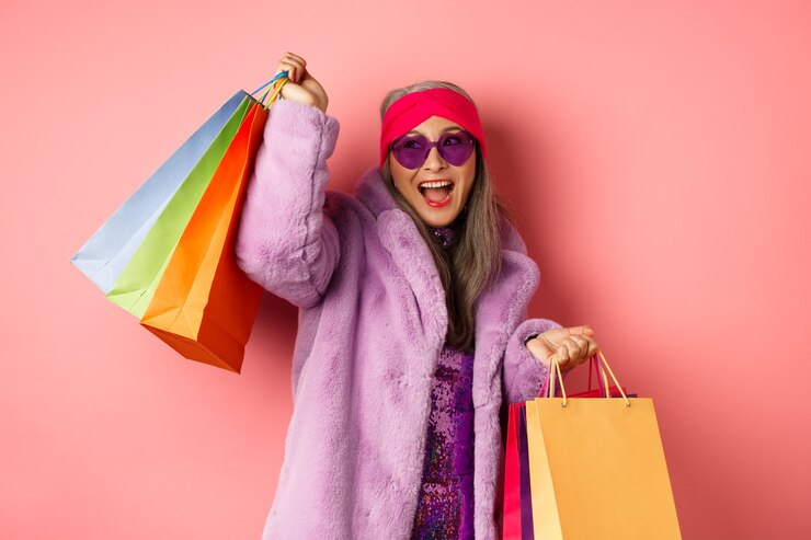Photo stylish asian senior woman going shopping, wearing trendy clothes and sunglasses, holding store bags and dancing happy, pink background