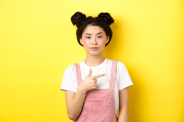 메이크업과 여름 옷을 입고 세련된 아시아 십대 소녀, 오른쪽 손가락을 가리키고 노란색 배경에 서 심각한 표정