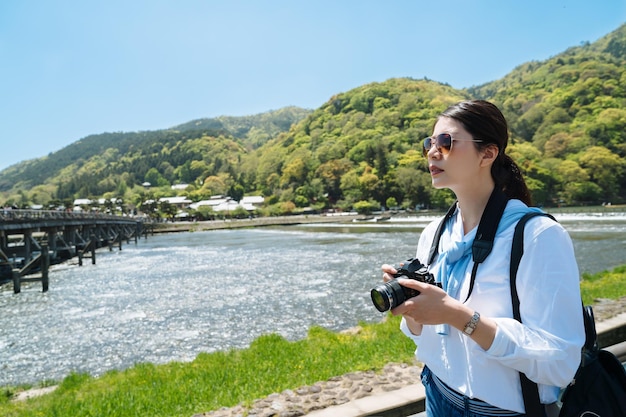 カメラを持ったスタイリッシュなアジア人女性が、川と山を背景に晴れた日に日本の嵐山の渡月橋の近くで遠くを見ている