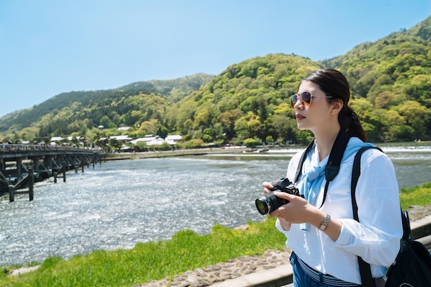 Stylish asian woman carrying camera is looking into distance near togetsu-kyo bridge in arashiyama, japan on a sunny day with river and mountains at background