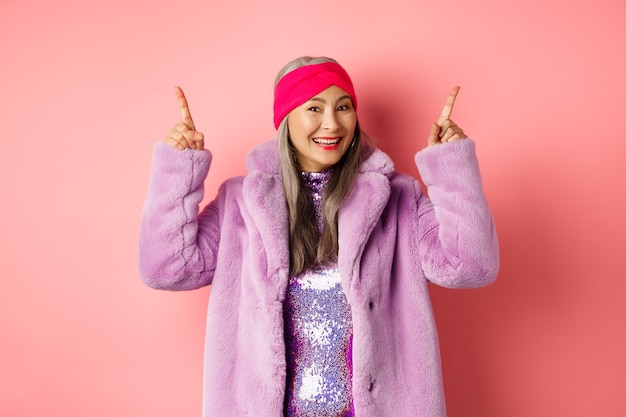 Stylish asian woman smiling happy, pointing fingers up and showing advertisement, wearing winter fake-fur coat and headband, pink background.