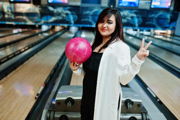 Stylish asian woman standing at bowling alley with ball at hand and show two fingers