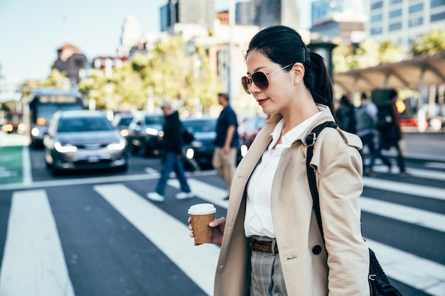 Stylish asian woman walking on crosswalk. young girl in sunglasses on pedestrian zebra crossing with cooffee cup in hand. people crowd in background and cars waiting for green traffic light.