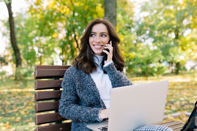 Stylish attractive businesswoman in grey pullover speaking on phone and working on laptop outside. She has short dark hair and big blue eyes, looking to side