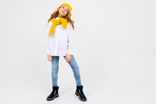 Стильная привлекательная мечтательная кавказская девушка в белой рубашке и синих джинсах и желтой шляпе в сапогах позирует с поднятой ногой на белой студии во всю длину