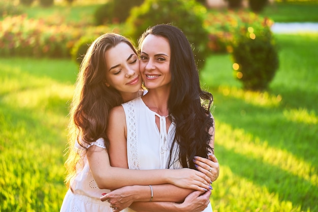 Stylish attractive smiling joyful happy mother and daughter hugging each other in a park outdoors at sunset at soft light