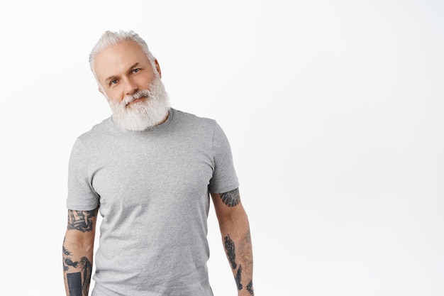 Stylish authentic senior man with tattoos and long beard, standing in casual grey t-shirt, looking happy and pleased at camera, smiling, standing over white background