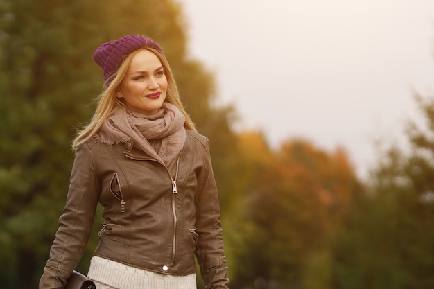 Stylish autumn. Fashionable pretty blond woman in cap, leather jacket and with handbag posing in autumn park, sunlight