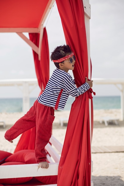 Stylish baby boy in a striped T-shirt and sunglasses stands the red beach sunbed