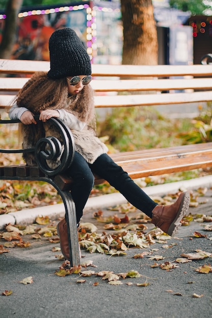 Stylish baby girl 4-5 year old wearing knitted hat, sunglasses, boots, fur coat sitting on the bench in park. Looking at camera. Autumn fall season.