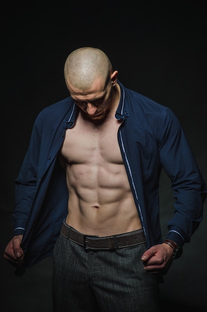 A stylish, bald young man stands with a naked torso on a dark background