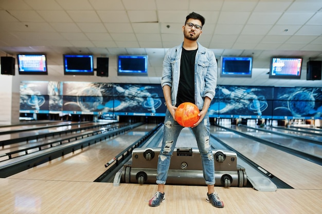 Stylish beard asian man in jeans jacket and glasses standing at bowling alley with ball at hand