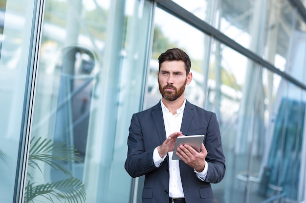 Stylish bearded businessman in formal business suit standing working with tablet in hands on background modern office building outside. Man using smartphone or uses mobile phone outdoors city street