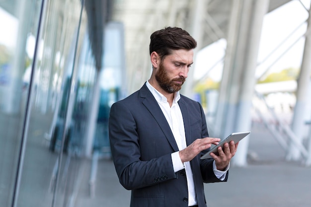 Stylish bearded businessman in formal business suit standing working with tablet in hands on background modern office building outside. Man using smartphone or uses mobile phone outdoors city street