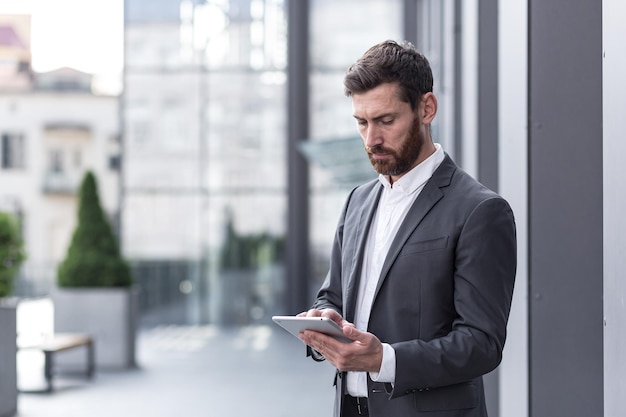 Stylish bearded businessman in formal business suit standing working with tablet in hands on background modern office building outside.