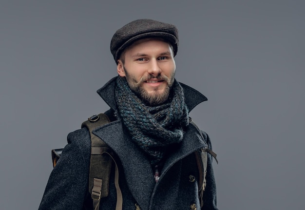 A stylish bearded hipster man dressed in a grey jacket holds a backpack.