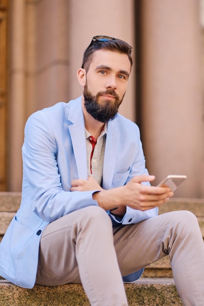 The stylish bearded male sits on a step and using smartphone.