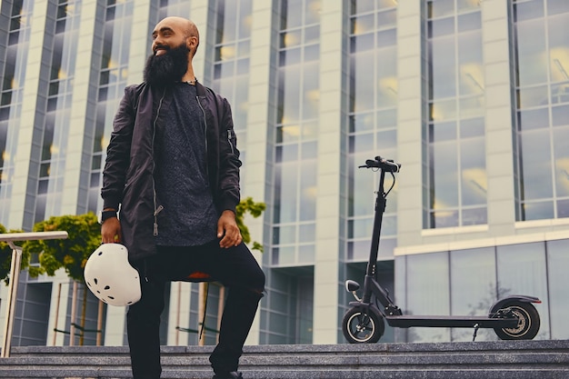 Стильный бородатый мужчина на ступеньке на фоне электрического скутера.