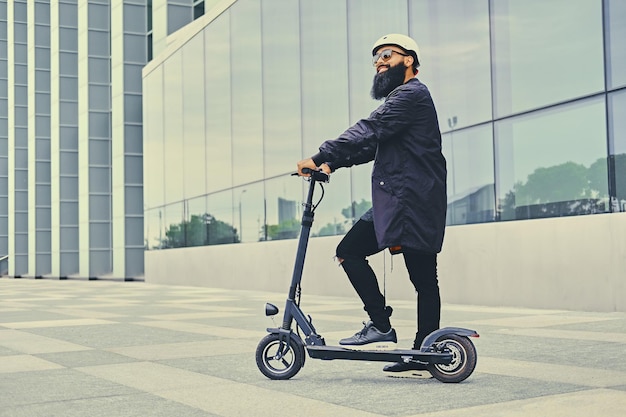 Stylish bearded male in sunglasses posing on electric scooter in over modern building background.