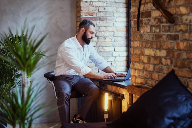 Il maschio barbuto alla moda lavora con un laptop in una stanza con interni soppalcati.