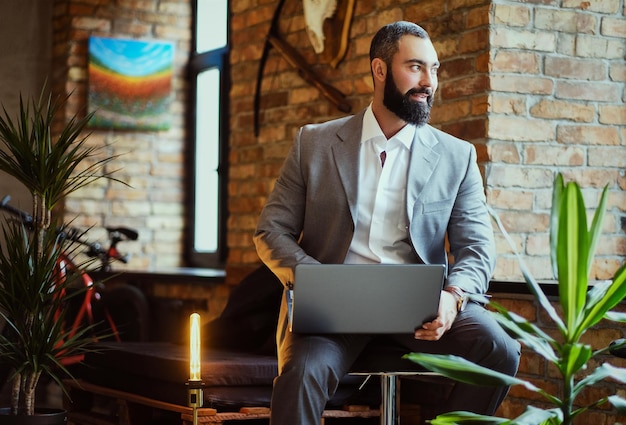 Il maschio barbuto alla moda lavora con un laptop in una stanza con interni soppalcati.