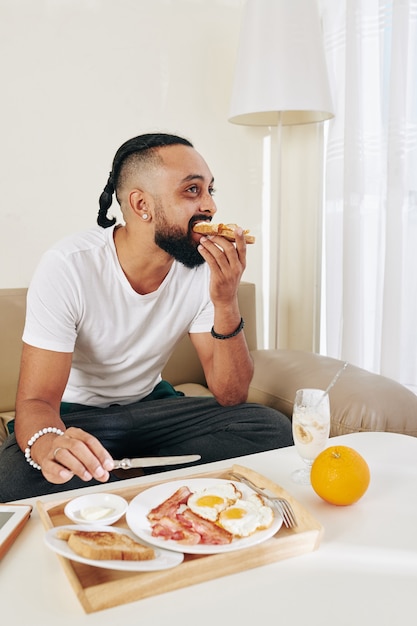 Stylish bearded man eating eggs with bacon, yogurt and sandwiches for breakfast at home