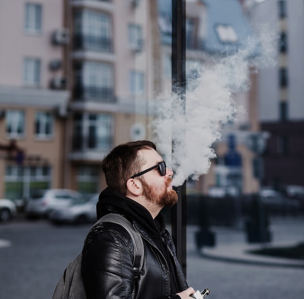 Elegante uomo barbuto con una giacca di pelle emette fumo dalla bocca