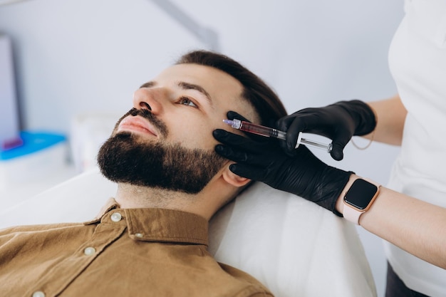 Стильный бородатый мужчина, посещающий эстетическую клинику, получающий наполнитель для губ крупным планом Привлекательный мужчина, делающий инъекции красоты в мужском спа-салоне на заднем плане