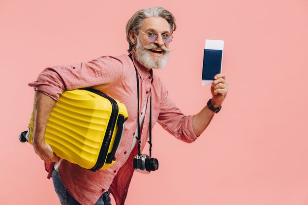 Фото Стильный бородатый мужчина с фотоаппаратом держит желтый чемодан и паспорт с билетом, улыбается и готовится к поездке.