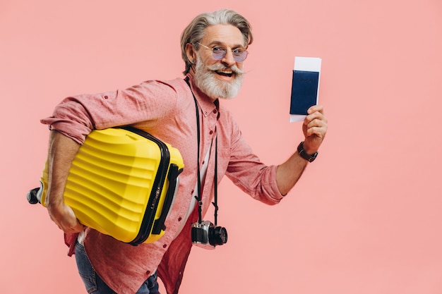 Elegante uomo barbuto con una macchina fotografica tiene una valigia gialla e un passaporto con un biglietto, sorride e si prepara per il viaggio.