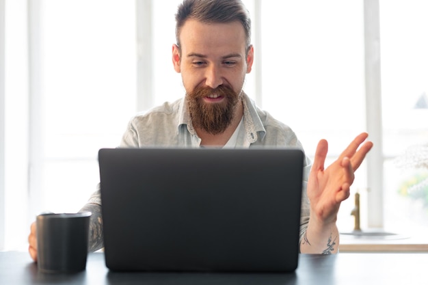 Uomo barbuto alla moda che lavora al computer portatile da casa seduto al tavolo