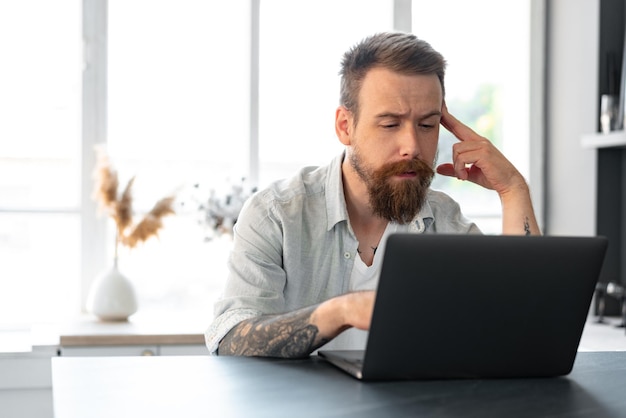 Stylish bearded man working on laptop from home sitting at the table