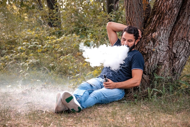 Stylish bearded smoker blows up a couple an e-cigarette on the forest floor. Electronic cigarette.