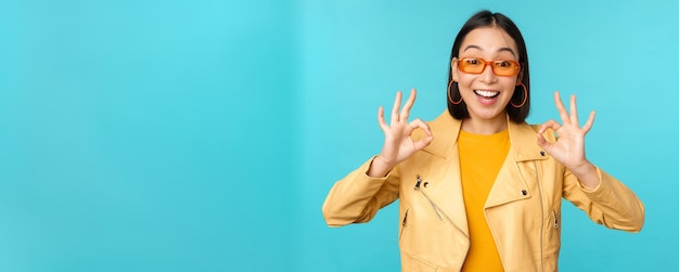 Stylish beautiful asian woman in sunglasses smiling amazed showing okay ok sign recommending smth supports excellent choice standing over blue background