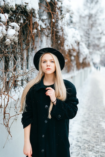 Stylish beautiful blonde girl in black clothes and hat posing near fence in the street