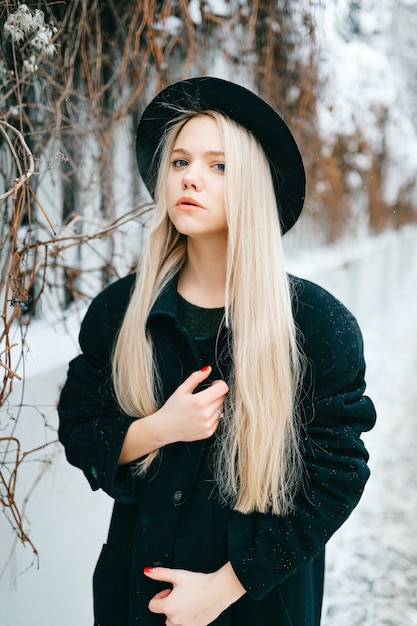 Stylish beautiful blonde girl in black clothes and hat posing near fence in the street