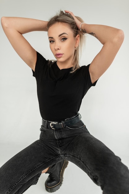 Photo stylish beautiful blonde girl in fashionable black clothes with a classic tshirt and black jeans sits and poses on a white background