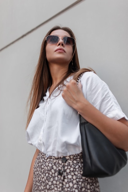 Stylish beautiful business girl model with vintage sunglasses\
in fashion casual summer clothes with a white shirt and a skirt\
with a bag walks on a gray background in the city