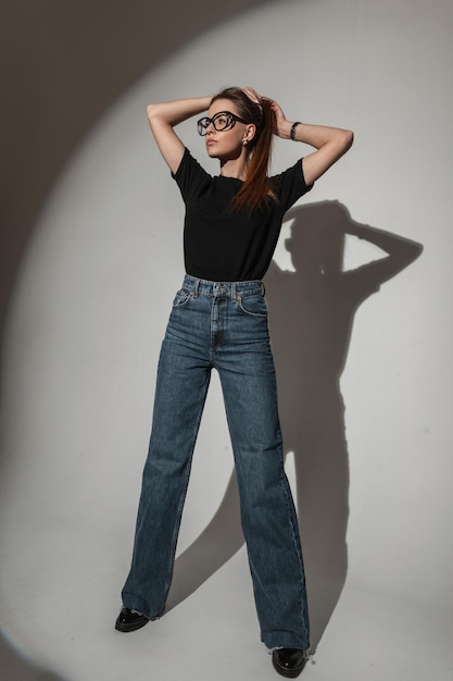 Стильная красивая гламурная молодая женщина в винтажных очках в черной футболке с синими модными джинсами в студии