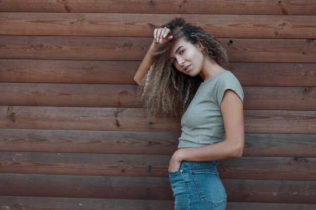 Stylish beautiful hipster woman with curly hair in vintage trendy jeans clothes posing near a wooden wall
