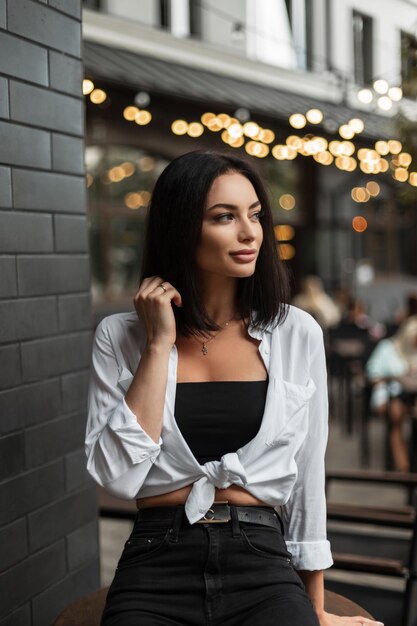 Stylish beautiful urban girl in fashionable casual clothes with a white shirt sits in the city near a black brick wall on a light bokeh background