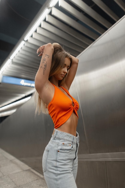 Stylish beautiful woman with a sexy body in a fashion orange top and vintage jeans on a metallic urban background in the subway