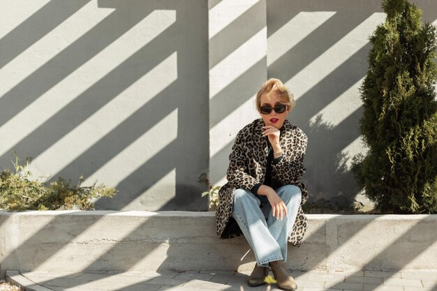 Stylish beautiful young blonde woman with sunglasses in a fashionable leopard coat with flared jeans and shoes sits on the street with sun light