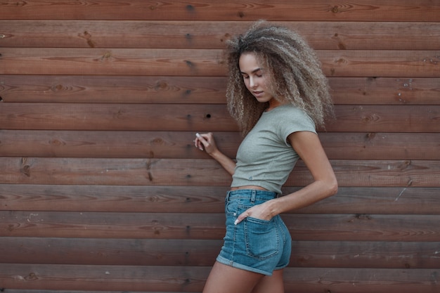 Stylish beautiful young curly hipster trendy woman in fashion denim clothes near a wooden wall