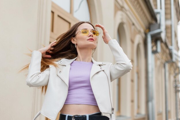 Elegante bella ragazza modello in abiti casual alla moda con giacca bianca in pelle e maglietta viola che cammina in città e si aggiusta i capelli