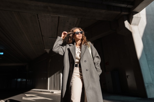 Stylish beautiful young girl with curly hair with sun glasses in a fashionable long coat walks in the city. Female model, urban style and beauty. Sunlight and shadow